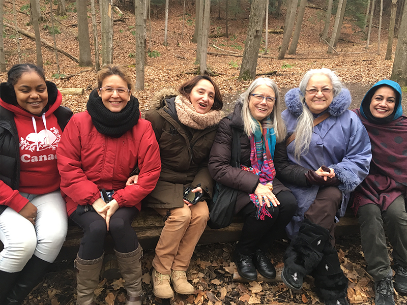 Six women sitting on a wooden log in a wooded area. They are leaning together and smiling at the camera, all wearing jackets, scarves, and other cold-weather outerwear. Behind them are evergreens and tree trunks emerging from a thick layer of autumn leaves.