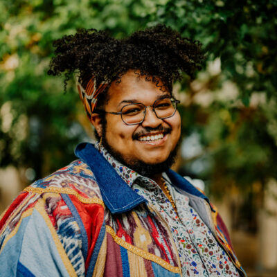 A head shot photo of Miguel Fenrich, a queer Black artist, smiling outside with greenery in the background.