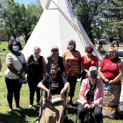 10 women pose in a group outside in front of a teepee. Most are wearing face masks; in the front row, two of the women are seated, one on a mobility aid.