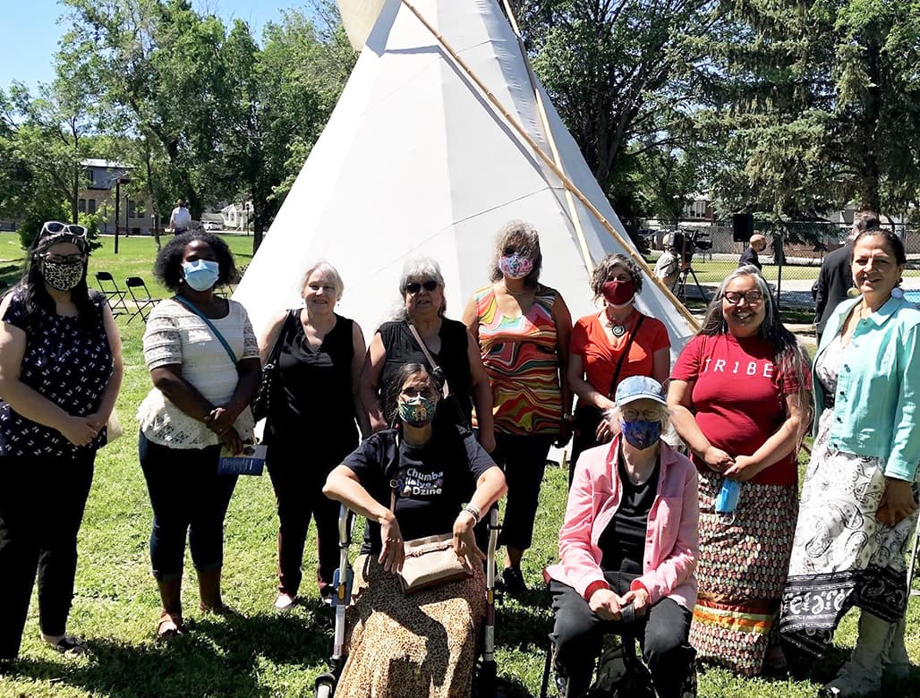 10 women pose in a group outside in front of a teepee. Most are wearing face masks; in the front row, two of the women are seated, one on a mobility aid.