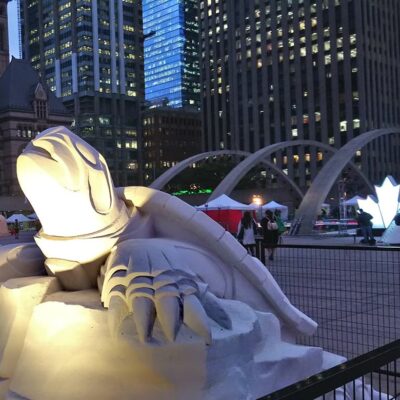 A photograph featuring a life-sized replica of a turtle statue in the foreground. It is made from white stone and is lit from beneath by a spotlight. Behind the statue are tents and teepees, as well as concrete arches and tall glass buildings. It is dusk.
