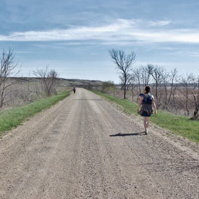 A woman walks down a gravel road with her back to the camera. In the distance another figure walks towards her. On either side of the road are bare trees, with a body of water on the right. The horizon shows rolling hills, and the sky is a bright blue with scattered clouds.