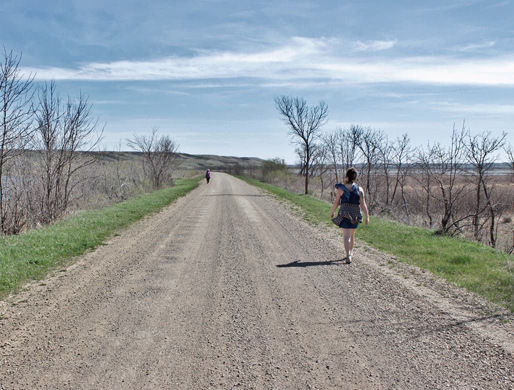 A woman walks down a gravel road with her back to the camera. In the distance another figure walks towards her. On either side of the road are bare trees, with a body of water on the right. The horizon shows rolling hills, and the sky is a bright blue with scattered clouds.