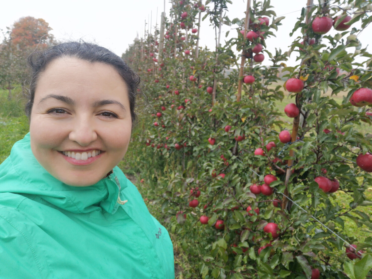 Image of a woman in a green windbreaker with brown hair that is pulled back. She is smiling at the camera next to a trellis of red flowers