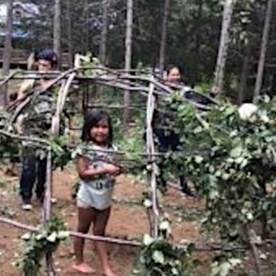 A young girl stands in the middle of the frame for a sweat lodge, constructed out of branches with leaves at the joints. Three women stand behind her, positioned around the sweat lodge frame.
