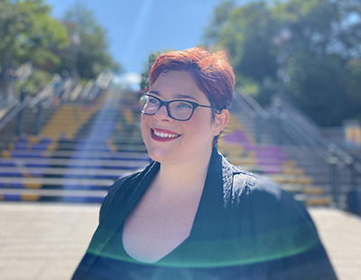 A plus-sized white woman stands outside in front of a set of concrete steps painted with a bright design. She has short red hair and glasses, and is wearing a black jacket and top. She is facing slightly to the left and smiling off camera.