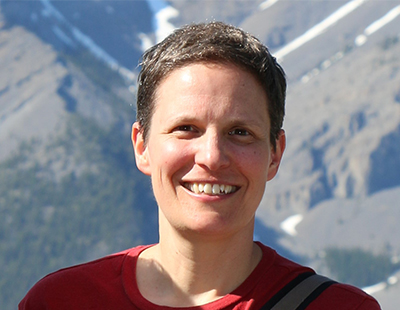 A white woman in her forties stands outside on a sunny day with a mountain in the background. She’s wearing a red t-shirt, has short brown hair and glasses, and is smiling at the camera.