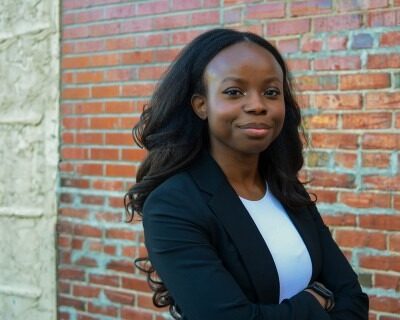 Photo of a young Black woman in a black and white shirt smiling confidently in front of a brick wall.