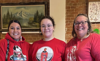 Three Algonquin Anishinaabekwes in red shirts, standing shoulder to shoulder.