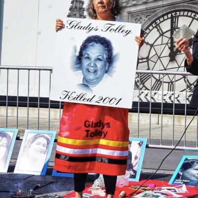 An Algonquin Anishinaabe/Kitigan Zibi First Nations woman in an orange ribbon skirt holds up a placard with a photo of Gladys Tolley that includes her name and the text "killed 2001"