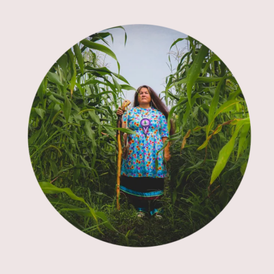 Wakerahkáhtste Louise McDonald Herne stands between two rows of tall cornstalks under a blue sky. She holds a staff and looks resolutely toward the camera.