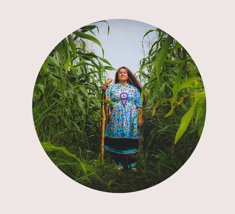 Wakerahkáhtste Louise McDonald Herne stands between two rows of tall cornstalks under a blue sky. She holds a staff and looks resolutely toward the camera.