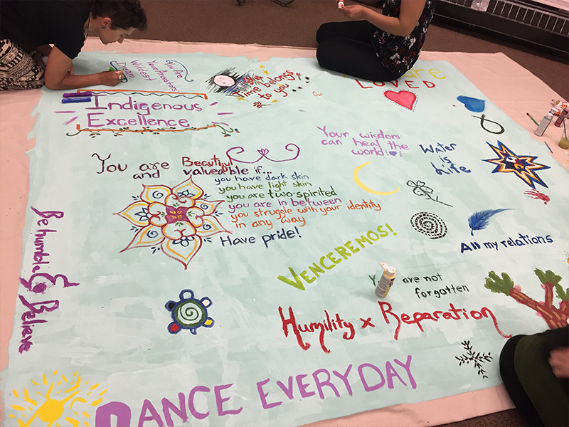 Photo of a large piece of white poster paper spread out on the floor. Three people are kneeling to paint on the paper, their upper bodies not visible. In colourful paint, messages have been written along with designs of a turtle, the medicine wheel, and abstract designs. Messages include “Indigenous excellence; Your wisdom can heal the world; You are not forgotten; Water is Life; All my relations”.