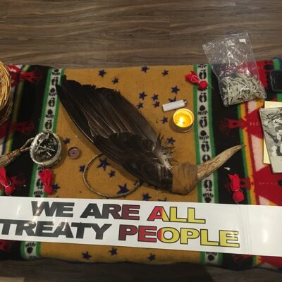 A collection of objects and medicines sits on top of a blanket spread over a table. There is a wing fan in the centre of the table, surrounded by red cloth bundles, candles, a book entitled “Poems of Rita Joe”, and smudging materials. A paper banner beneath the items reads “we are all treaty people”.