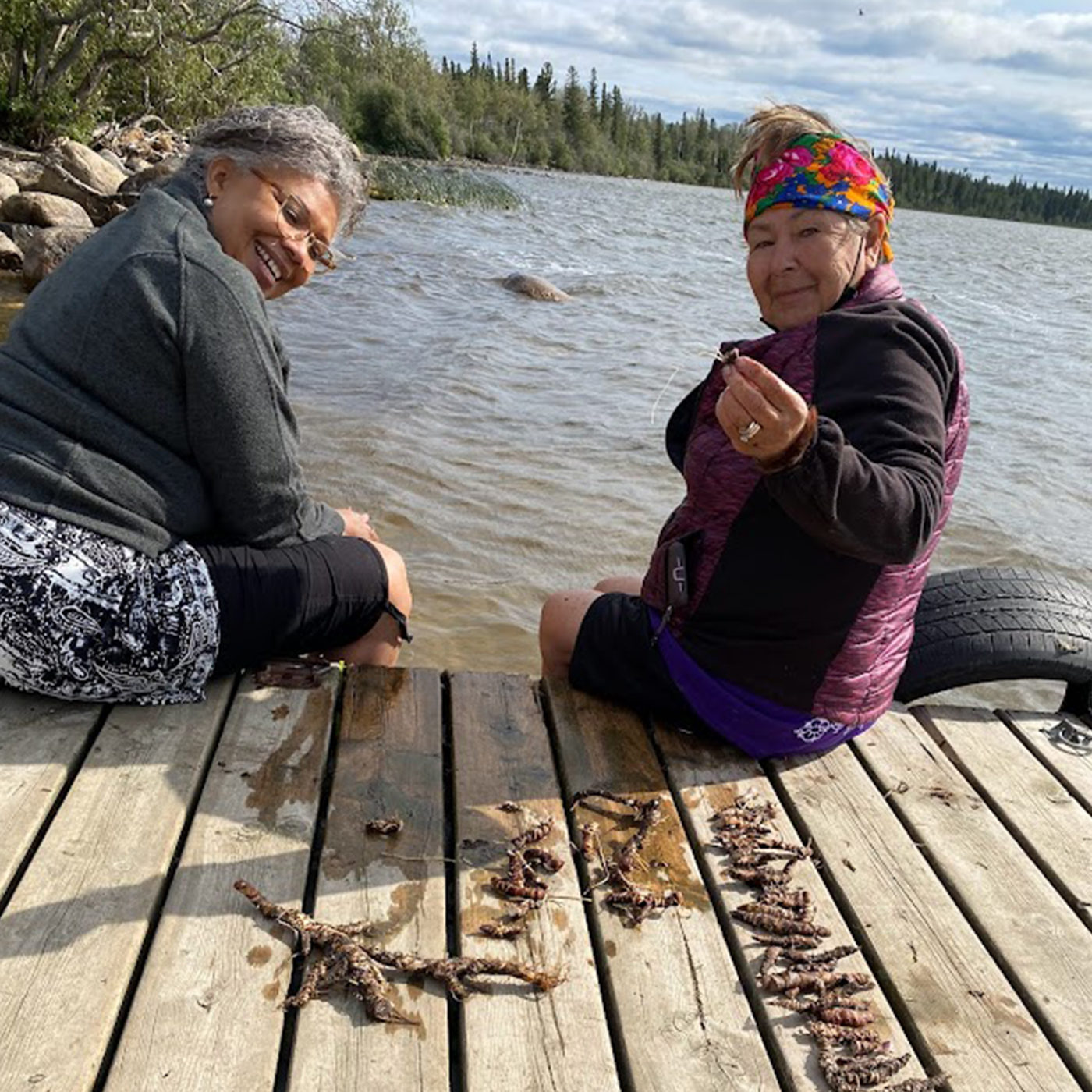 A Black and an Indigenous woman are seated on the edge of a wooden dock, with their legs in the water. They are looking over their shoulders towards the camera and smiling, with water and trees behind them. On the right, the Indigenous woman is reaching out to show the camera a piece of a root. On the dock next to them are more pieces of washed roots.