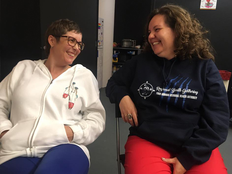 Two women looking at each other and laughing; they are both seated and turned slightly towards each other. The woman on the left wears a white hoodie with a feather graphic; she has short hair and glasses. The woman on the right wears a black hoodie with a logo; her hair is shoulder-length and curly.