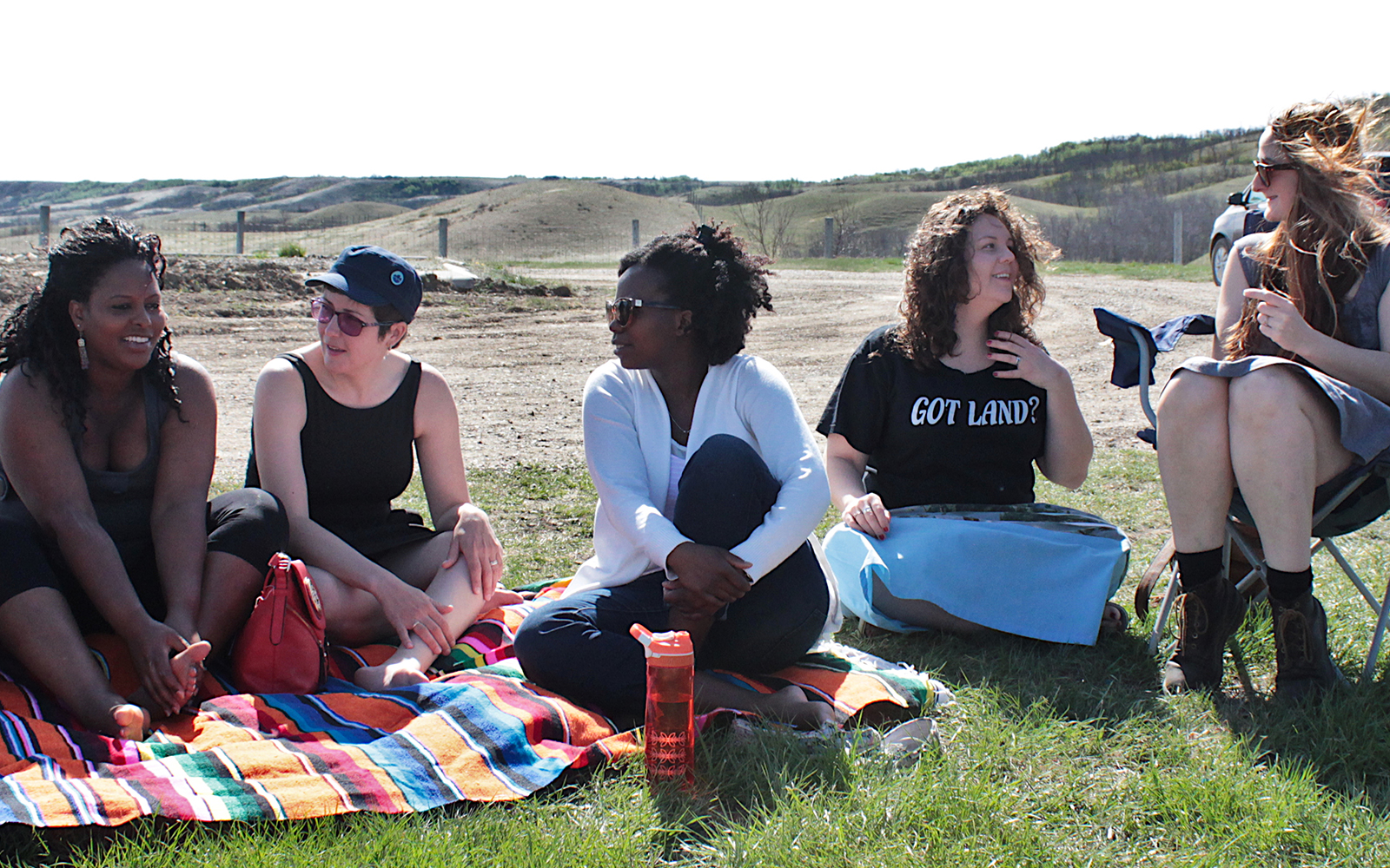 Five women are sitting outside with rolling green hills and sky behind them. Three of the women sit on a blanket on the left of the image, leaning towards each other in conversation. Two more women on the right - one seated in a folding chair, the other on the ground - are also talking while the wind blows their hair.