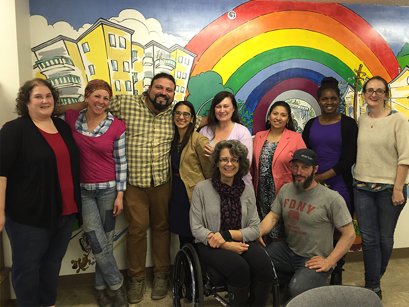 A photograph of 10 people standing against a wall with a bright mural painted on it. In the back row, either people stand facing the camera, smiling, some with their arms around each others shoulders. In the front row a white man in a baseball cap kneels next to a white woman in a wheelchair. The mural behind them depicts bright yellow buildings against a colourful rainbow.