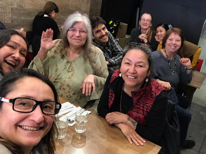 Eight people out for dinner lean in for a selfie. In the foreground, two women lean in to look at the camera with bright smiles, while next to them a woman in a green blouse waves. To her right is another women wearing a black top and red patterned vest, smiling. At the table behind them, three more women and a man lean in to the photo with wide smiles.