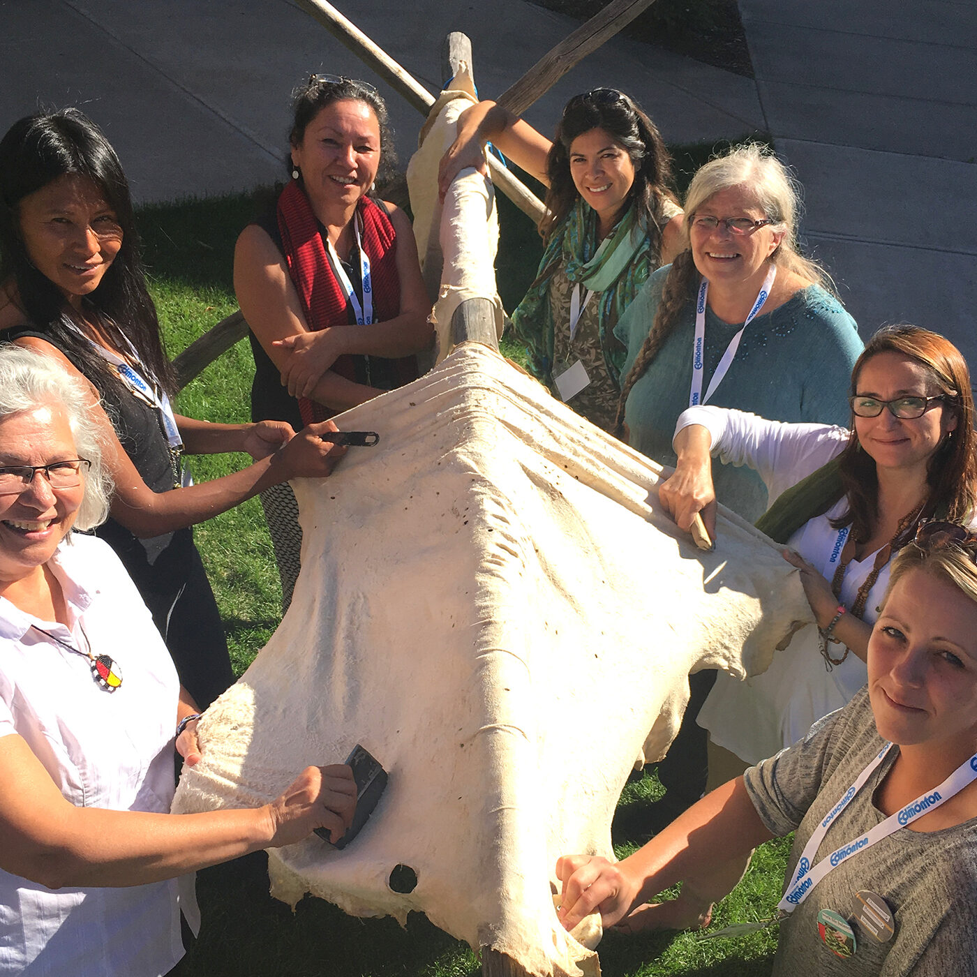 Seven women stand in a semi-circle around a moose hide stretched over a wooden frame. They are outside, and behind them is grass and a cement walkway. Some are scraping the hide, others are just watching. They are all looking at the camera and smiling in the sunlight.