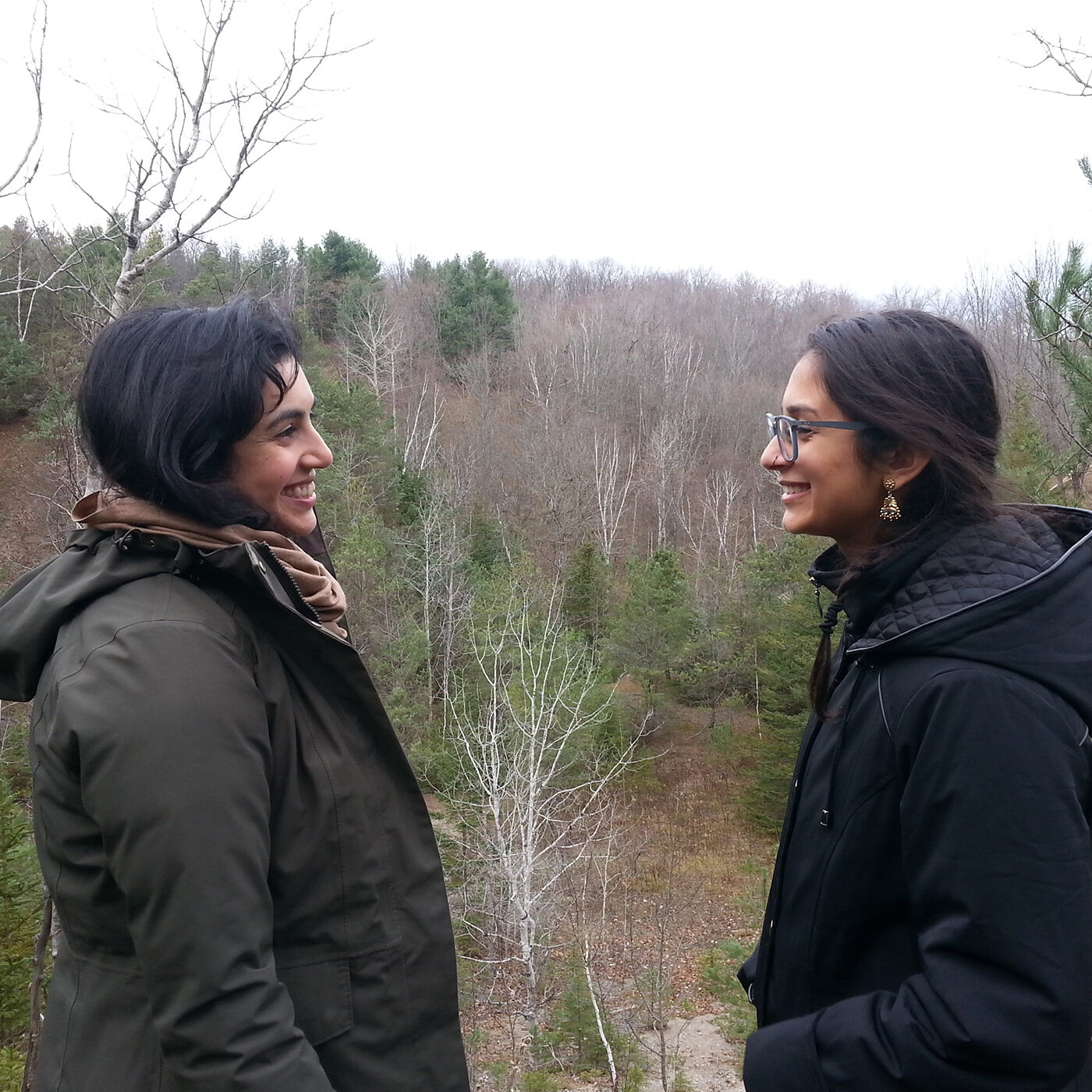 Two women stand facing each other against a wide landscape of bare trees and evergreens. Both have long dark hair and light brown skin, and both are wearing warm jackets and smiling at each other.