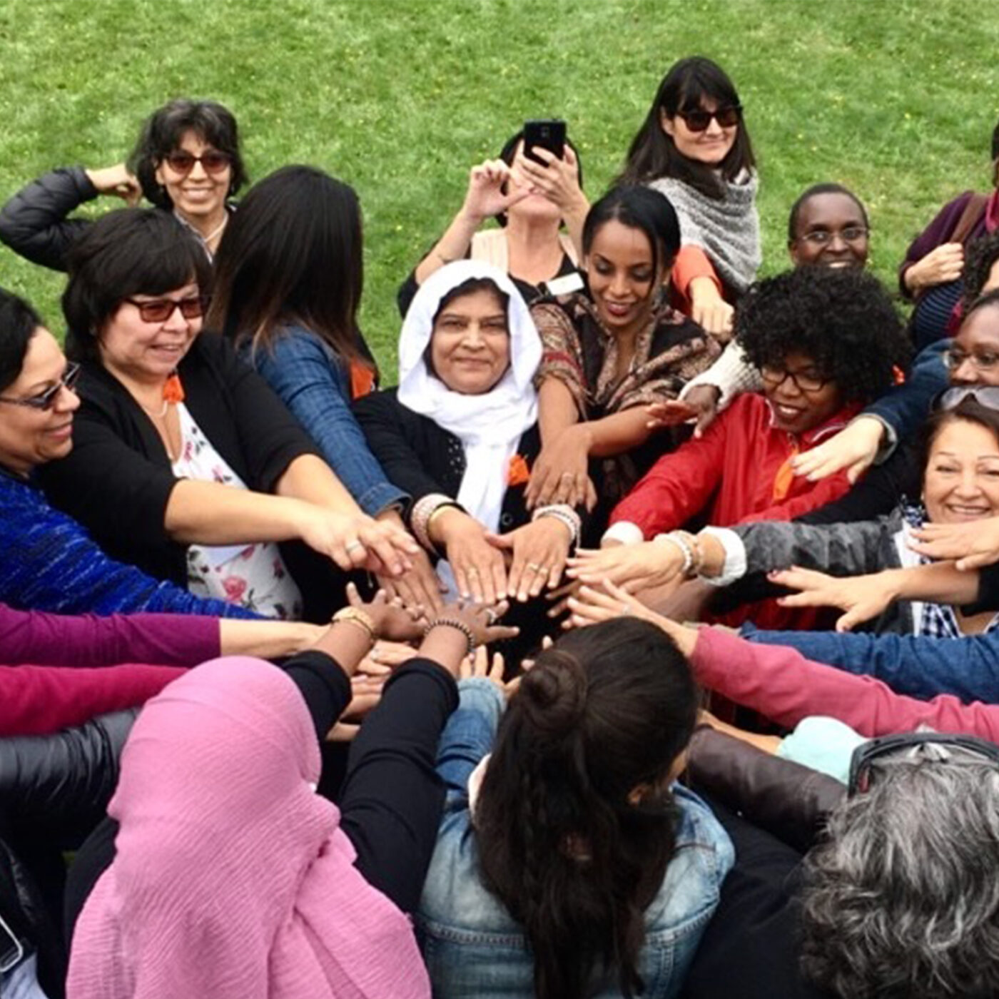 A large group of Black, Brown, white, and Indigenous women stand close together in a circle, smiling as their arms reach into the centre. Some are looking at the camera, others are taking pictures with their own phones.
