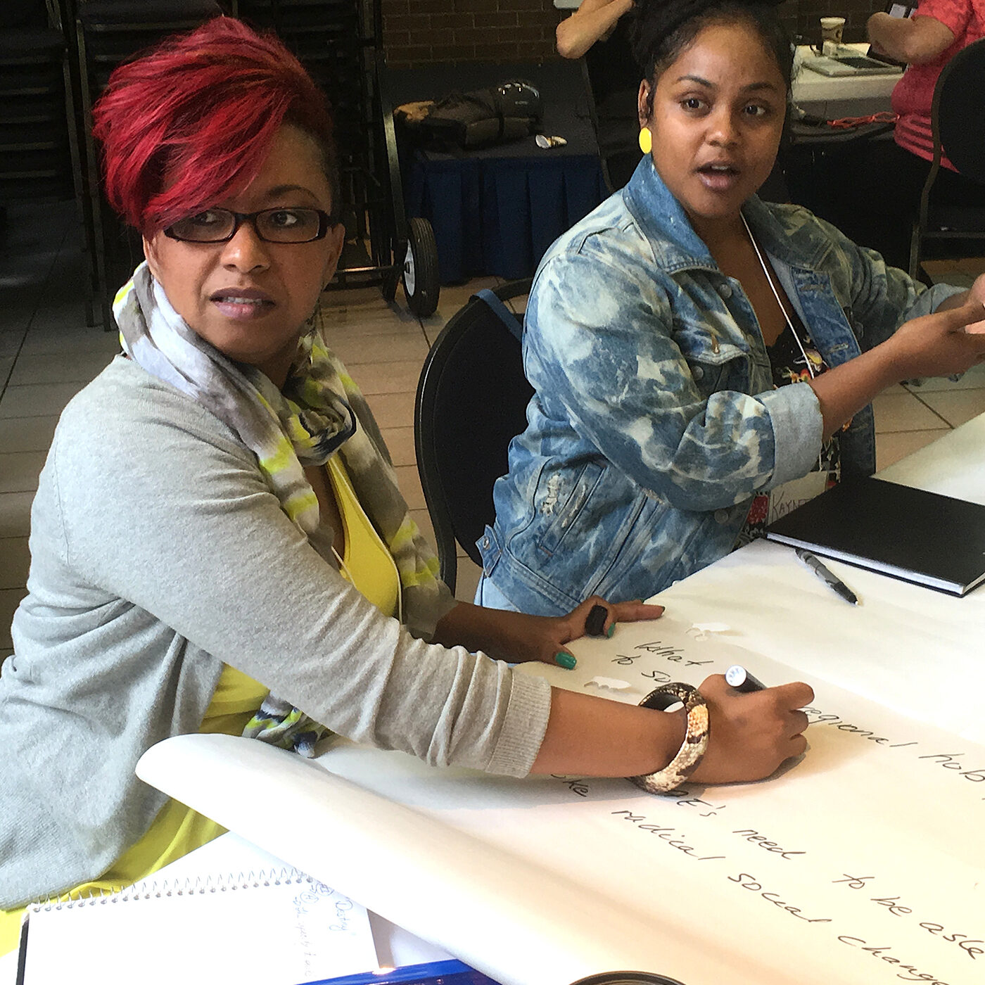 Two Black women are seated at a table, with a piece of flip-chart paper in front of them, both looking off-camera to the left. The woman on the right looks like she’s speaking; she’s wearing a blue denim jacket, yellow earrings, and her hair is pulled back. She’s gesturing as she speaks. The woman on the right is writing on the flip-chart paper; she’s wearing a grey cardigan over a yellow top. Her short red hair is swept to the side and she is wearing glasses.