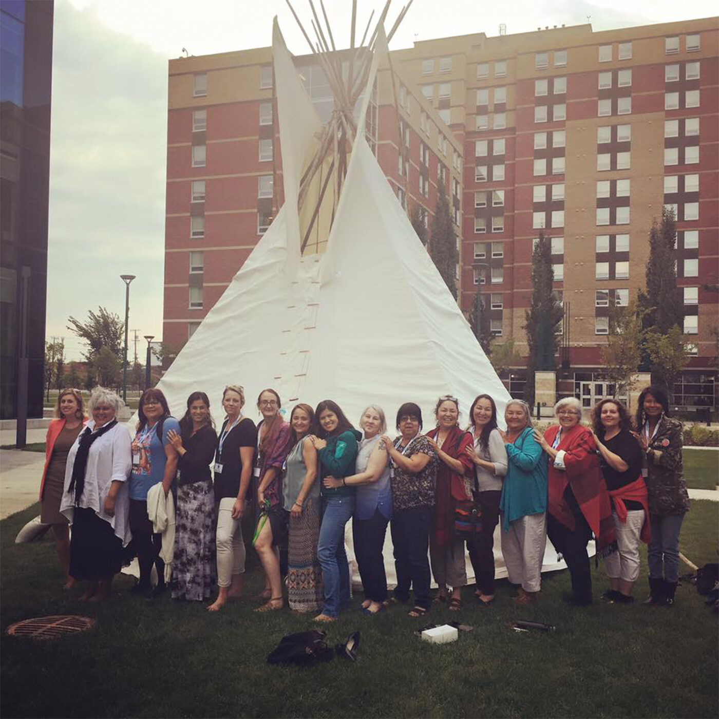 Sixteen women stand in a line in front of a teepee made of white fabric over a wooden frame. They are turned to the left and smiling towards the camera. Many have their hands resting on the shoulders of the woman in front of them, and in front of them some shoes and purses are scattered on the grass. Behind them is a multi-story red brick building and a bright grey sky.